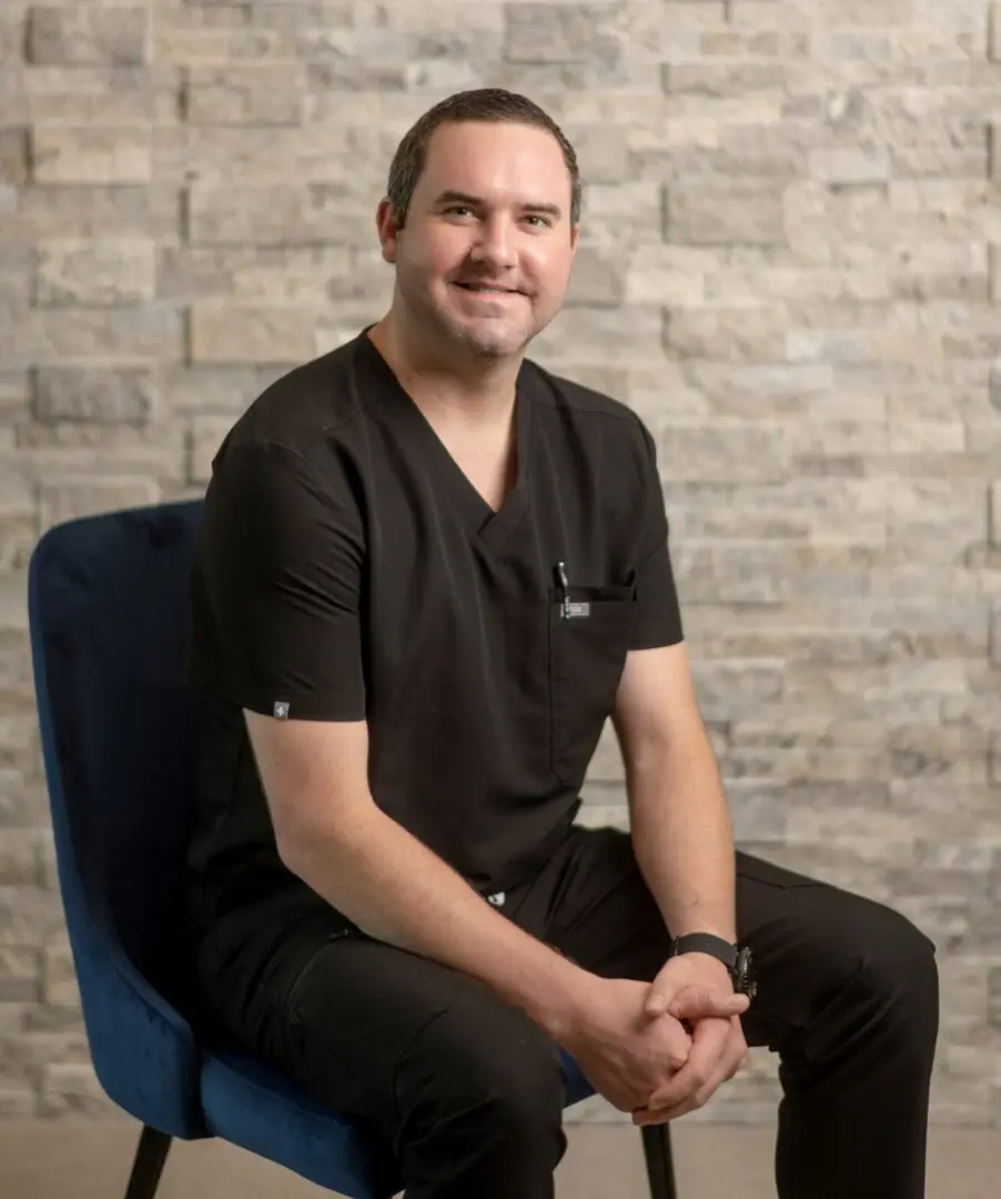 Dr-Evan-Hamilton-Photo-for-Staff-Page-1283x1536 Man sitting on chair against brick wall.