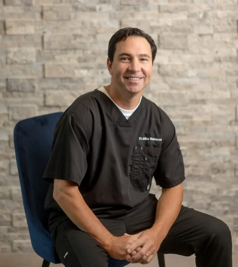 Dr.-Mike-Blanscet-Photo-for-Staff-Page-1371x1536 Smiling man sitting on a blue chair.